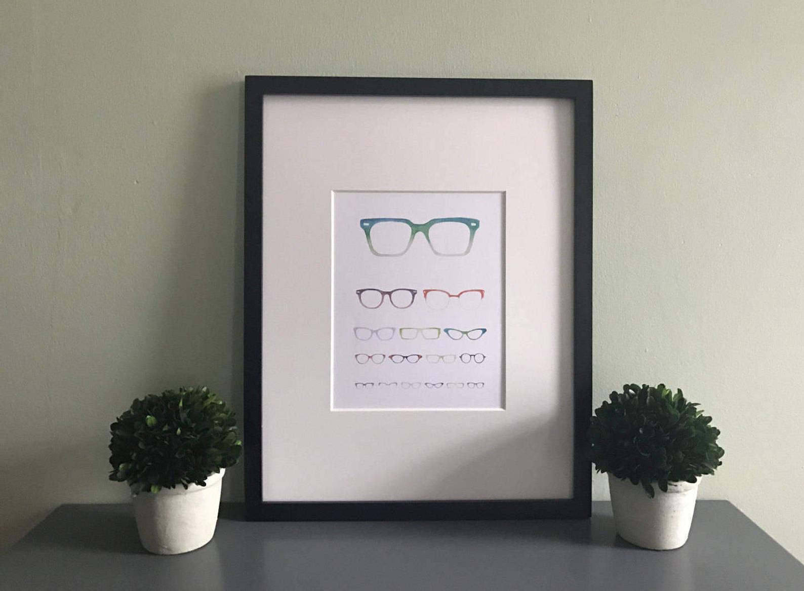 A print of an eye chart made up of varying sizes and styles of glasses. Shown in a black frame.