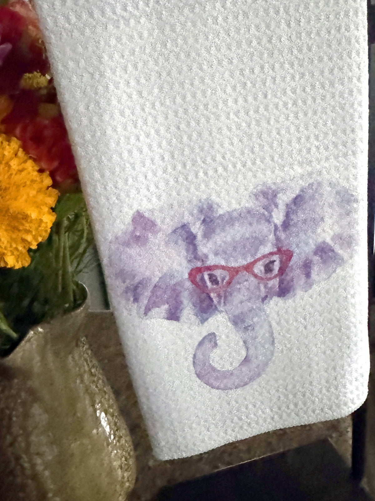 A white textured kitchen towel with a purple elephant graphic wearing red glasses, hanging on a black stand next to a vase of colorful flowers.