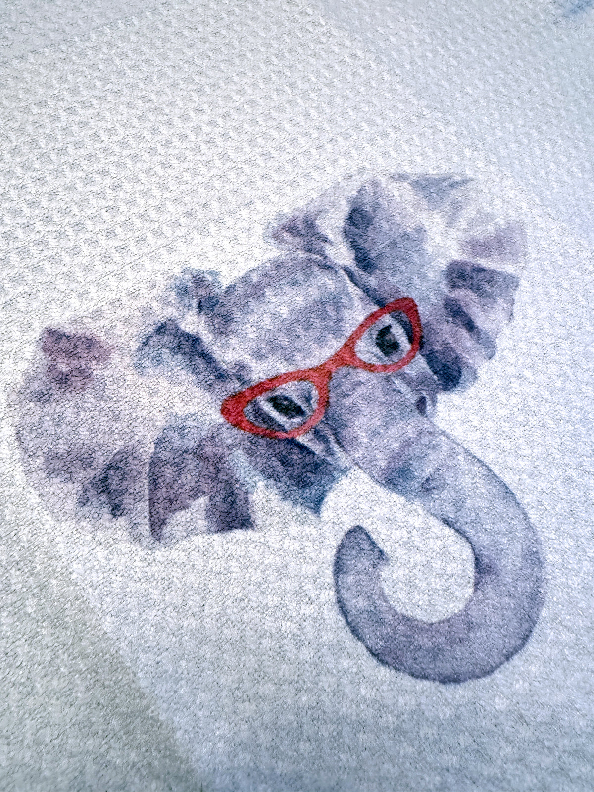 A close-up of a gray elephant graphic wearing red glasses on a textured white fabric background.