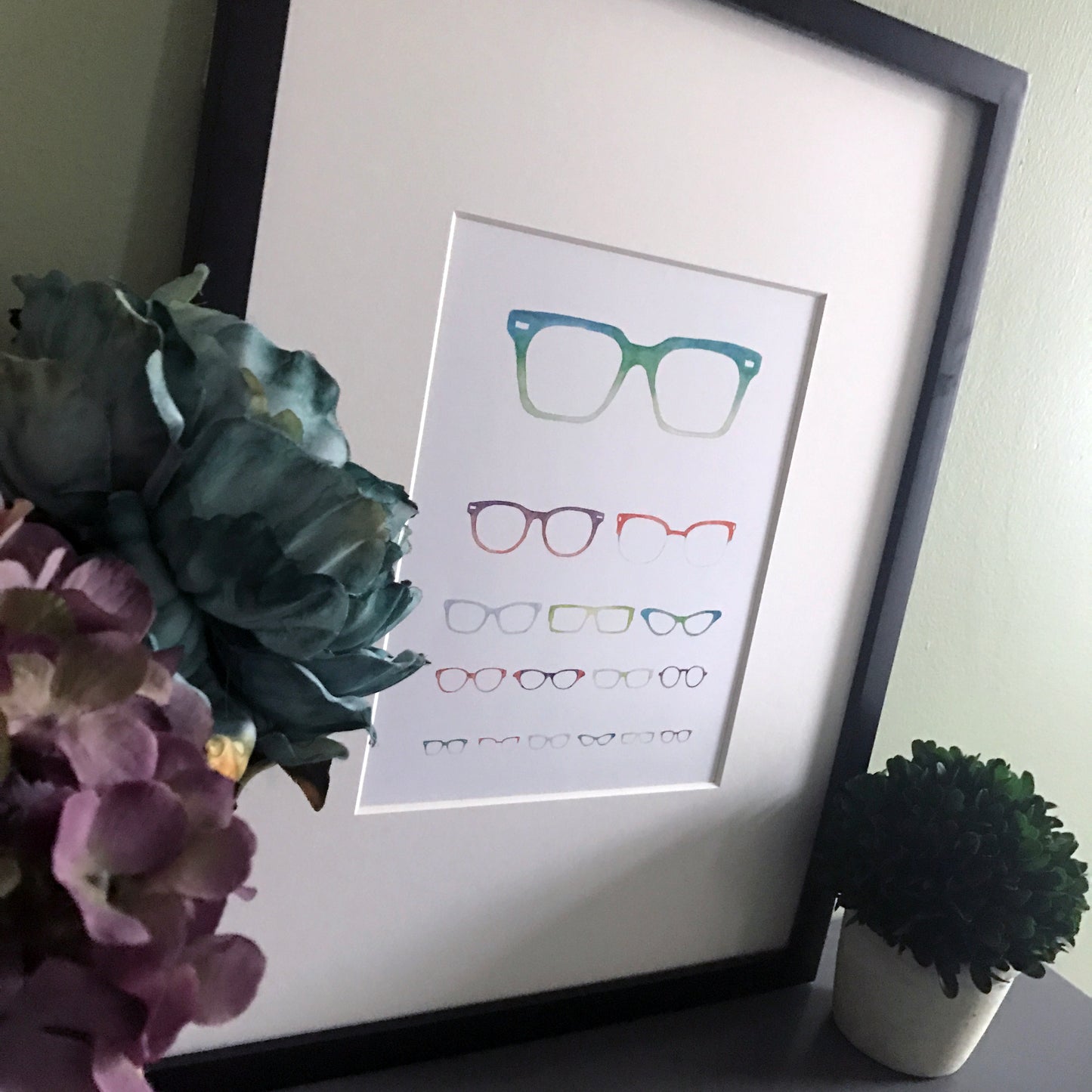 A print of an eye chart made up of varying sizes and styles of glasses. Shown displayed in a black frame.