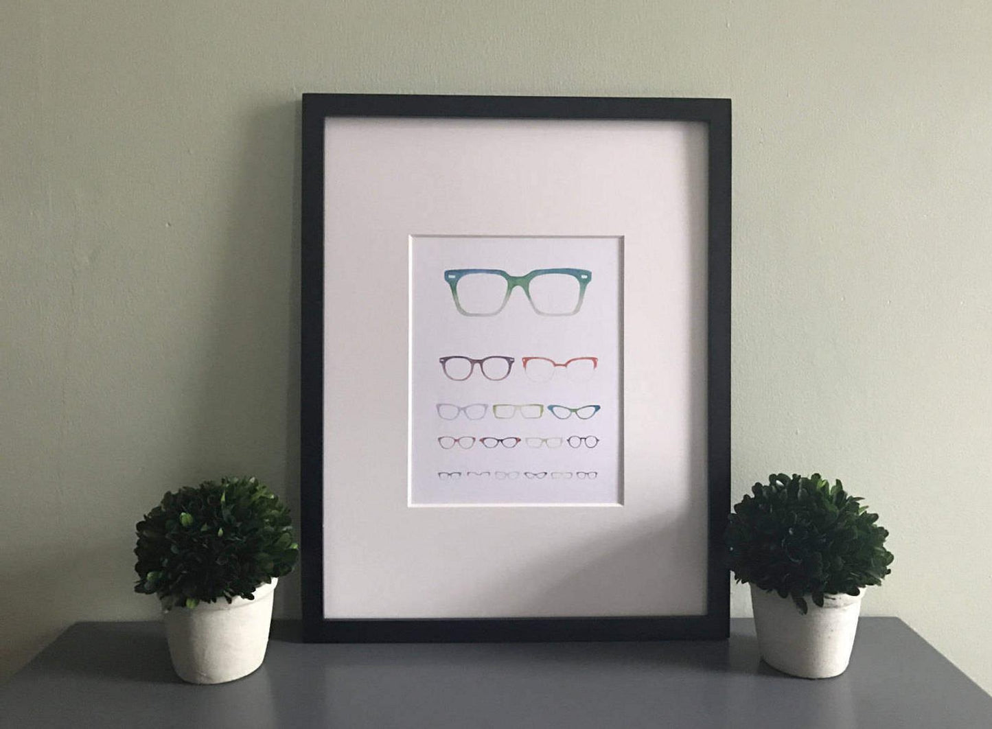 A print of an eye chart made up of varying sizes and styles of glasses. Shown in a black frame.