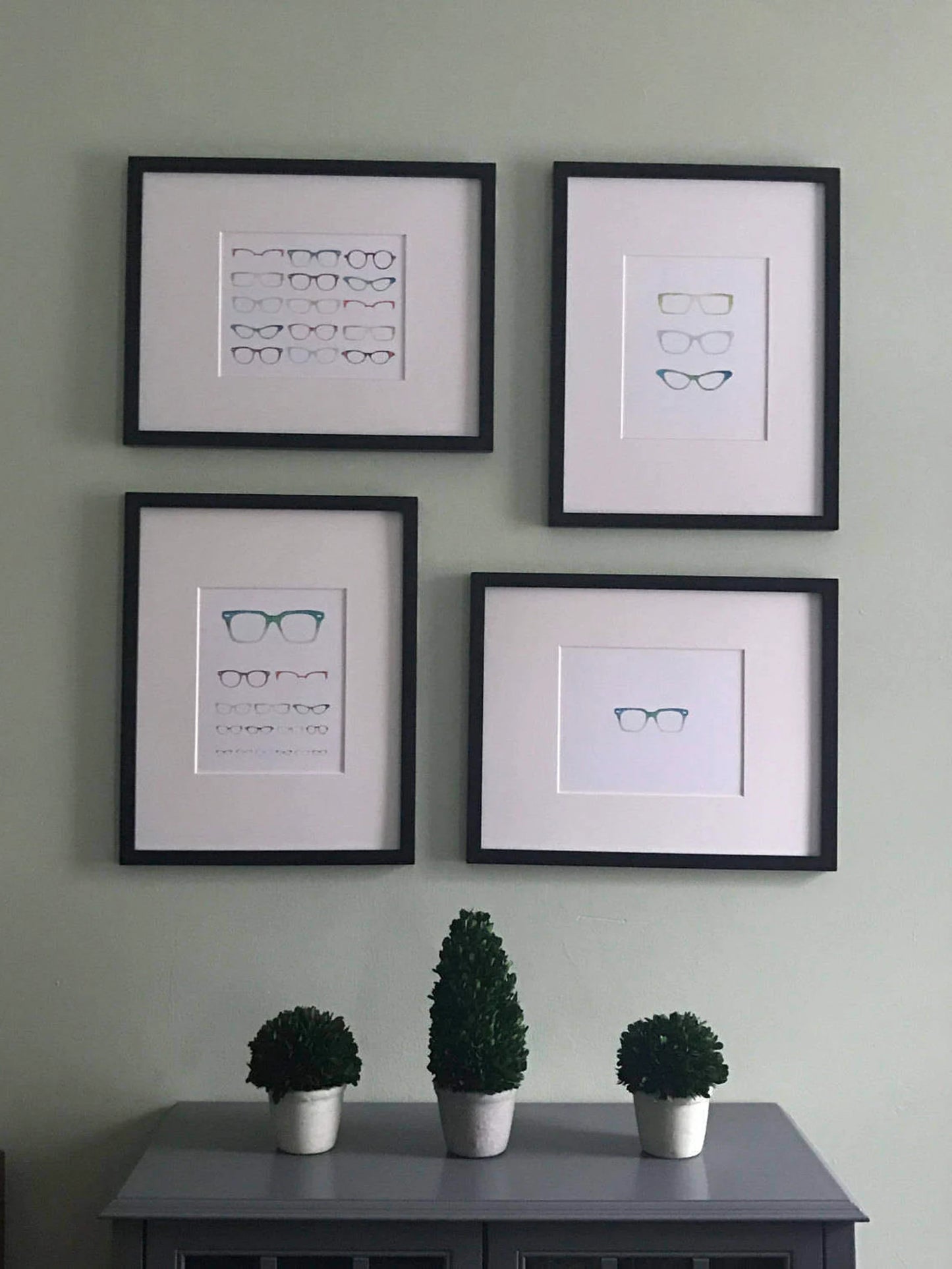 A print of an eye chart made up of varying sizes and styles of glasses. Shown displayed on a wall with other prints.